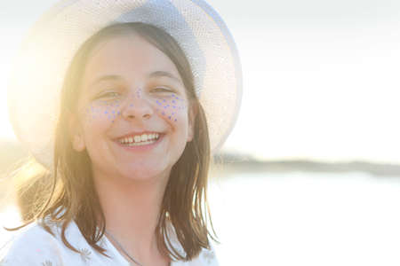 Happy playful female teenager with painted blue freckles wearing white hat looking at camera while...