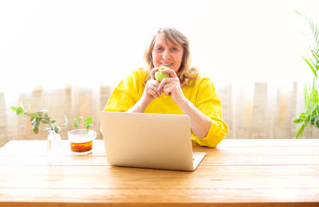 Positive middle aged female in casual wear sitting at wooden table with cup of tea and apple and browsing laptop while spending morning time at homeの写真素材