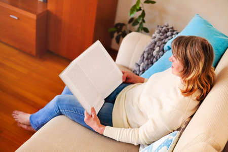 From above side view of calm mature female in casual clothes sitting on comfortable sofa and reading...
