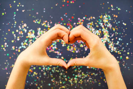 Top view of crop anonymous female with bright manicure showing heart gesture with fingers over dark...