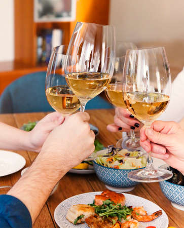Group of crop anonymous friends clinking glasses with wine while gathering around table with delicious dishes during home dinnerの写真素材
