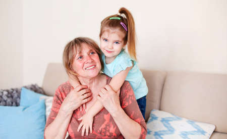 Smiling middle aged grandmother and little granddaughter hugging and looking at camera while sitting...