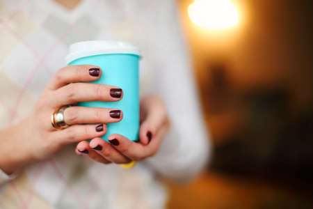 Crop anonymous female in warm sweater with dark manicure holding disposable cup of coffee while resting at wooden tableの写真素材