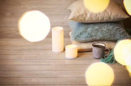 Mug with hot coffee placed on wooden floor near stacked cozy cushions and glowing candles in...
