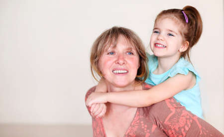 Smiling middle aged grandmother and little granddaughter hugging and looking at camera while sitting...