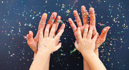 Top view of crop woman with manicure and little child holding hands on navy blue surface sprinkled with colorful confettiの写真素材