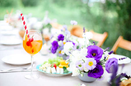 Glass of delicious Aperol spritz drink with straw placed on banquet table with appetizers and fresh flowers in gardenの写真素材