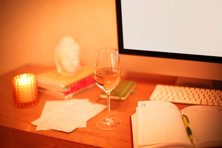 Glass of wine placed on table near notebook and computer monitor with blank screen in evening in home officeの写真素材