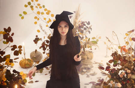 Young woman in witch costume looking at camera and using wand while standing on background of dry...