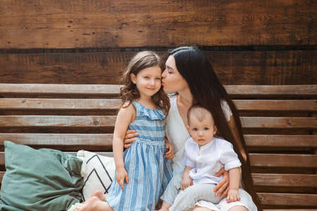 Young delighted mother sitting in armchair with adorable little siblings on terrace of country house and relaxing together during weekendの写真素材