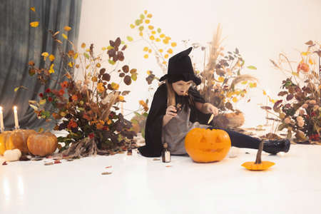 Little cute girl with long hair standing on scene in black dress and hat with autumn decorations on background during Halloweenの写真素材