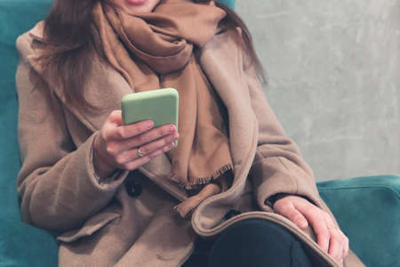 Crop female in warm brown coat and scarf interacting with smartphone while sitting in soft armchair in corridor of contemporary high rise buildingの写真素材