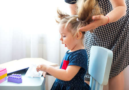 Crop woman doing ponytails for adorable little girl sitting at table and playing with toys during weekend at homeの写真素材