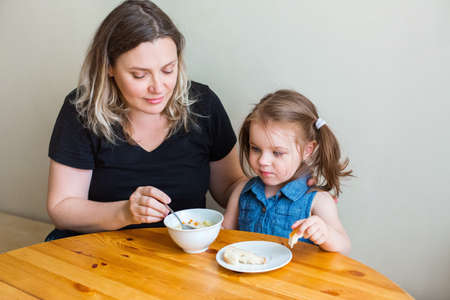 Content grandmother sitting at table in kitchen and feeding little granddaughter with tasty lunchの写真素材