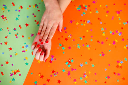 Top view of crop anonymous female with bright manicure showing heart gesture with fingers over orange and green background with colorful confettiの写真素材