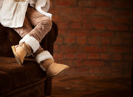 Faceless little girl in warm clothes and footwear sitting on brown couch