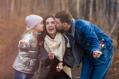 Cheerful young family with daughter dressed in warm clothes with sparklers in hands embracing and...