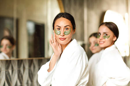 Company of smiling women in bathrobes and eye patches standing near mirror in bathroom and looking at camera while doing daily skincare routineの写真素材