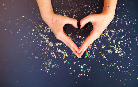 Top view of crop anonymous female with bright manicure showing heart gesture with fingers over dark...