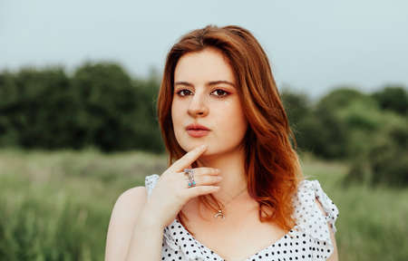 Side view of young female touching ginger hair and resting while sitting in grass on summer day in...
