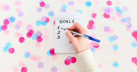 Female hand with pen writing New Year goals in empty notebook on white background with confetti