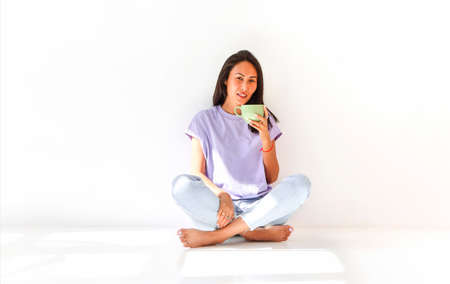Carefree ethnic female in domestic wear sitting on floor in room and enjoying fresh tea during...