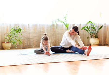 Mother and little daughter sitting on mat and doing exercises while warming up and training together at homeの写真素材