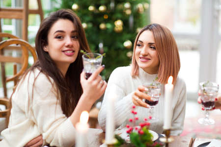 Content young women in casual wear with glasses of cocktails with red berries at festive table during Christmas holidayの写真素材