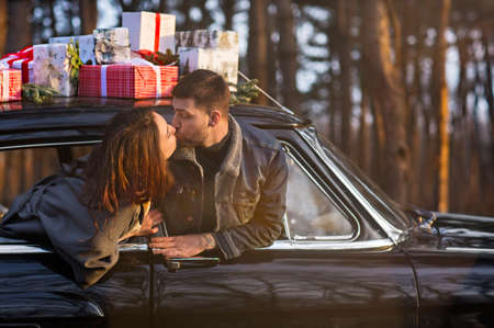 Loving man and woman sticking out of retro car windows and kissing with Christmas gifts on roof in...