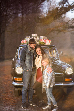 Smiling adult man and woman with little girl in warm clothes blowing snow standing at vintage car with Christmas presentsの写真素材