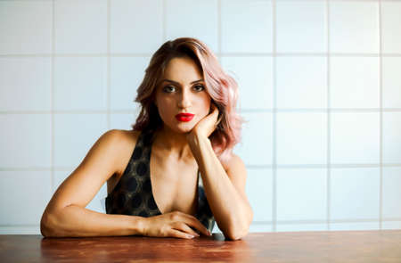 Charming female with red lips and in elegant dress sitting at wooden table while leaning on hand and looking at cameraの写真素材