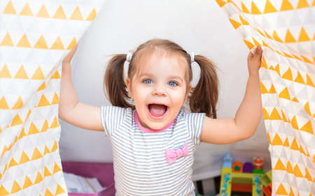 Cute little girl in casual clothes laughing at camera with various toys at home