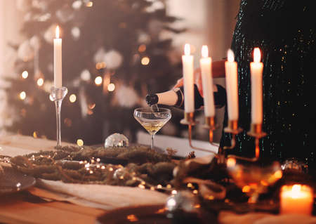 Unrecognizable female filling wineglass with champagne while standing near banquet table with burning candles during Christmas party at homeの写真素材