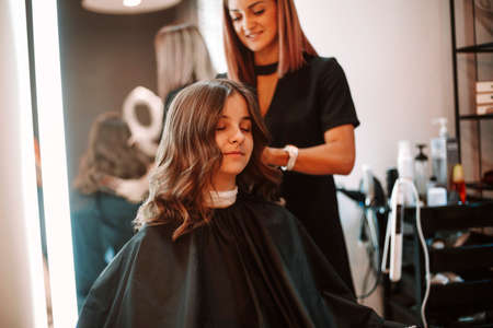 Young female customer with dark wavy hair getting professional hairdressing from positive hairstylist in modern beauty salonの写真素材