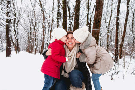 Little girls in outerwear kissing happy woman in cheeks while resting in winter forest together
