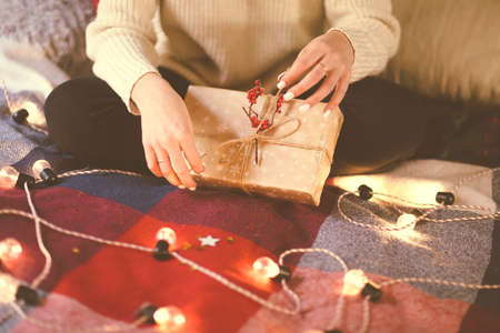 Crop woman in casual outfit packing Christmas gift box with cord and red berries while sitting on bed covered with plaid and decorated with light garlandの写真素材