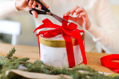 Young girl in white sweater packing round present box tied with bright red ribbon during holiday...