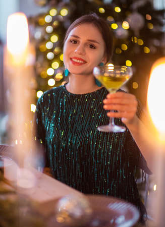 Female in shiny dark green party dress holding glass with champagne while standing against blurred background with light garlandsの写真素材