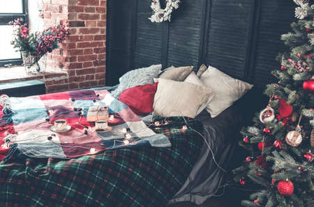 Cups of coffee and gift box placed on cozy bed with colorful checkered blanket and various pillows decorated with light garland and starsの写真素材