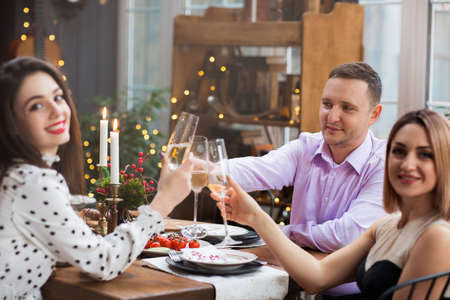 Cheerful young friends sitting at festive table and clinking glasses with champagne while having...