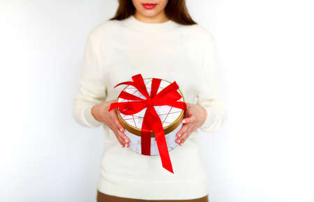 Unrecognizable girl in white sweater holding round present box tied with bright red ribbon during holiday celebrationの写真素材