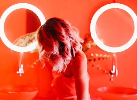 Back view of elegant female standing in contemporary red bathroom with sinks and illuminated mirrorsの写真素材