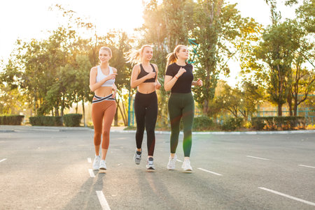 Full body young sportswomen smiling and running on asphalt parking lot on sunny day in parkの写真素材