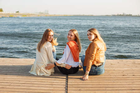 Back view of cheerful young women in casual clothes sitting on quay near water and looking at camera over shouldersの写真素材
