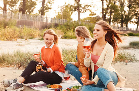 Cheerful young girlfriends drinking wine and eating bruschettas while having picnic on summer weekend day in parkの写真素材
