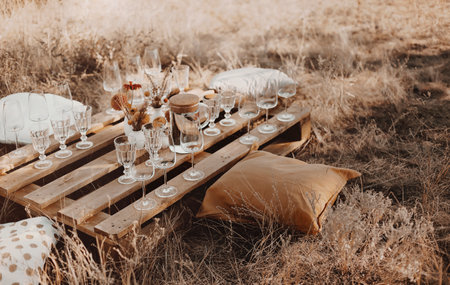 Set of elegant glassware placed on plank wooden table with floral arrangement on grassy meadow during festive autumn picnicの写真素材