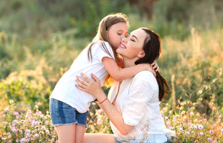 Tender daughter kissing delighted mother in cheek while resting in blooming meadow in summerの写真素材