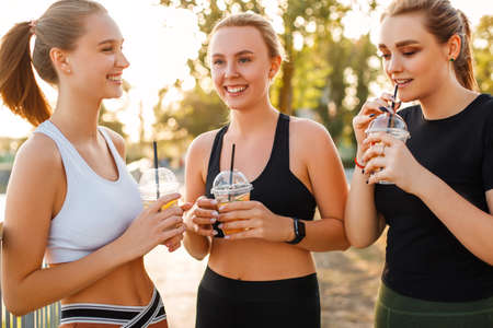 Young female in sportswear smiling and looking away while enjoying fresh healthy beverage near girlfriendsの写真素材