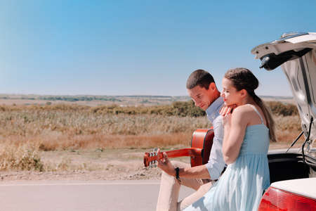 Happy young couple playing guitar and enjoying time together while sitting on open trunk of car parked on country road during summer tripの写真素材