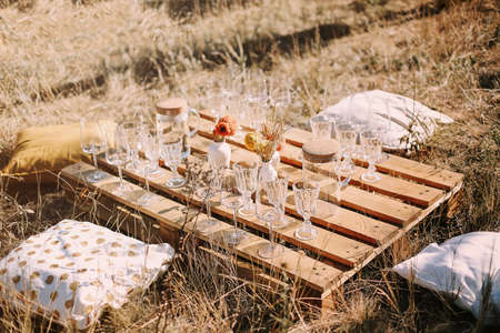 Set of elegant glassware placed on plank wooden table with floral arrangement on grassy meadow during festive autumn picnicの写真素材